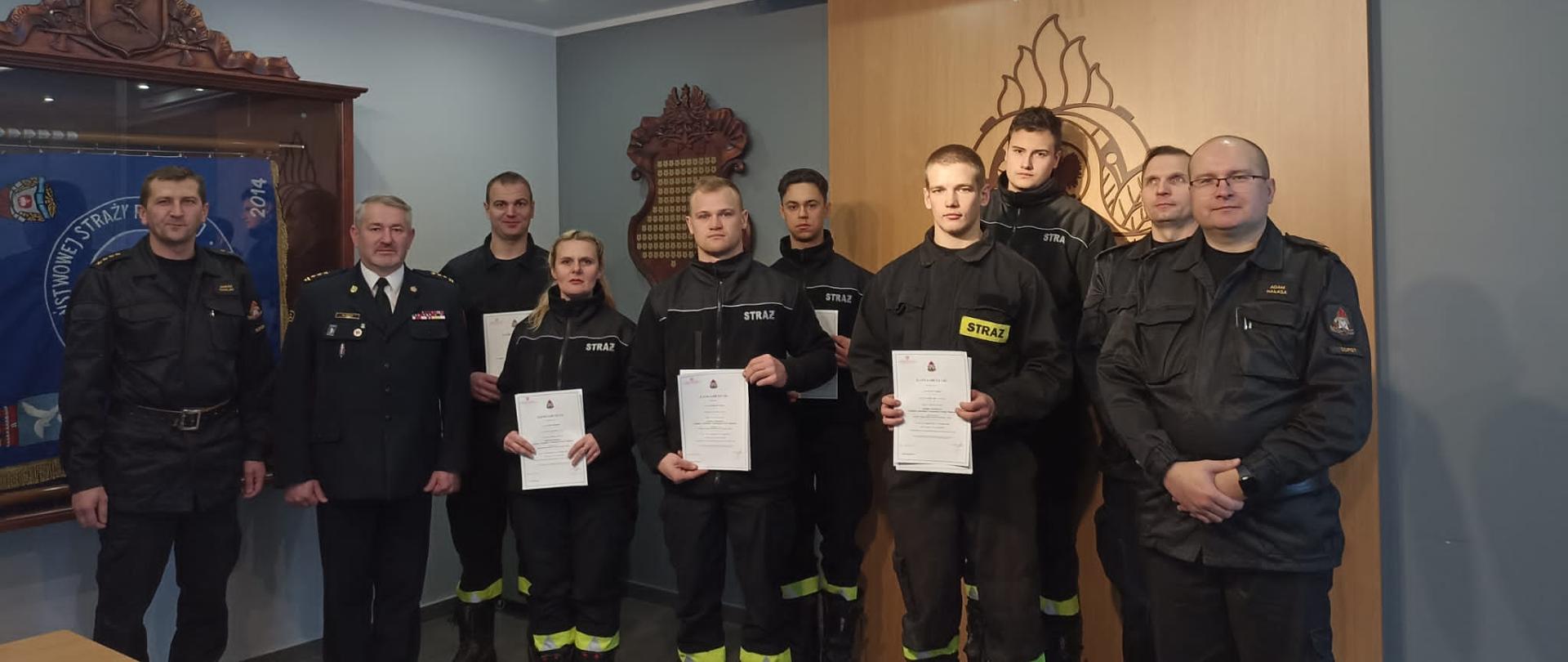 Zdjęcie grupowe druhów OSP trzymających odebrane zaświadczenia wraz z komendantem miejskim i członkami komisji w śwo=ietlicy
