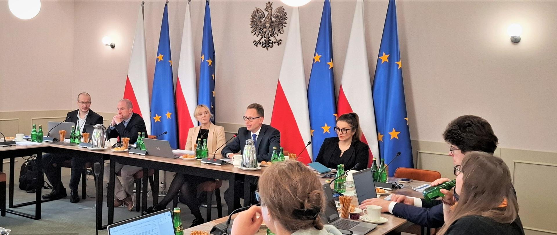 Przy stole konfernecyjnym siedzi grupa urzędników, w tle godło Polski i flagi. 