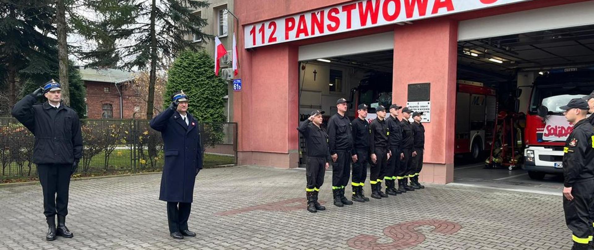 Komendant powiatowy PSP w Żyrardowie st. bryg. Mariusz Tymoszewicz wraz z dowódcą zmiany salutują przed budynkiem komendy. Obok stoją strażacy podziału bojowego w dwóch rzędach na przeciwko siebie.