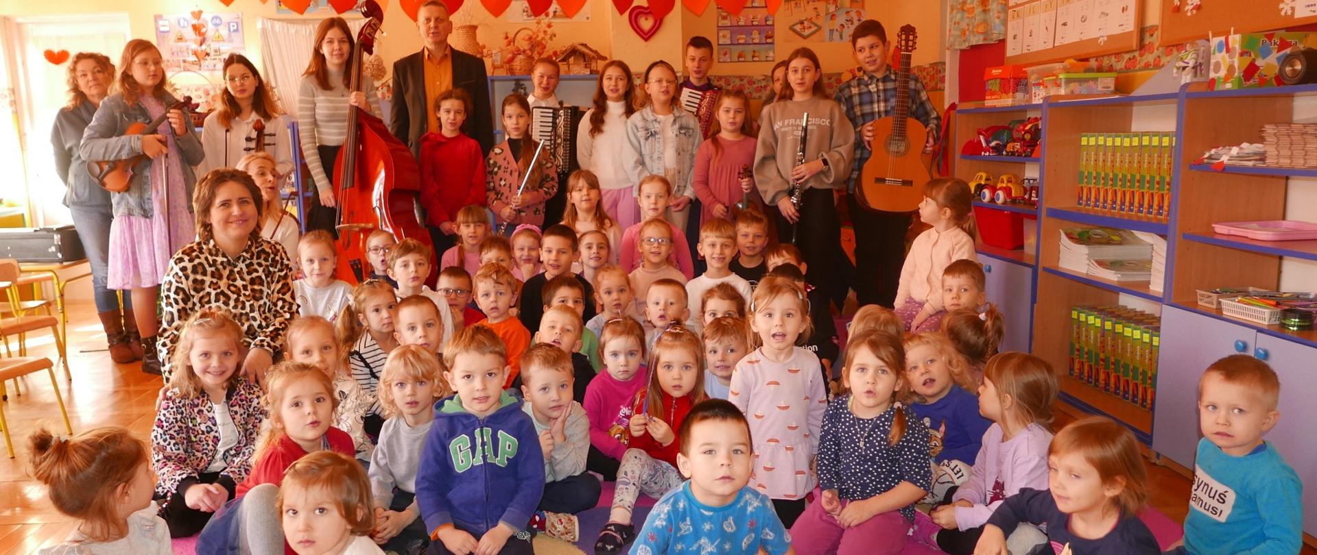 zdjęcie grupowe. Na dywanie siedzą przedszkolaki. W tle stoją uczniowie szkoły muzycznej, którzy trzymają instrumenty takie jak: flet poprzeczny, skrzypce, gitarę, akordeon.
