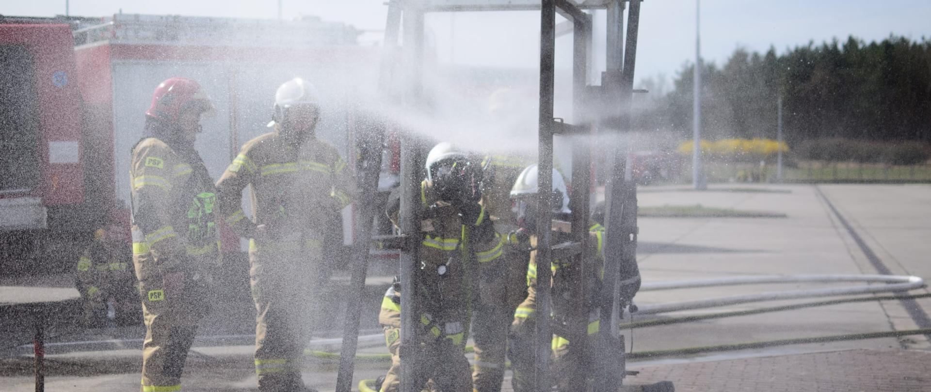 Strażacy ćwiczą na trenażerze do otwierania drzwi podawania wody przy wejściu do pożaru. 