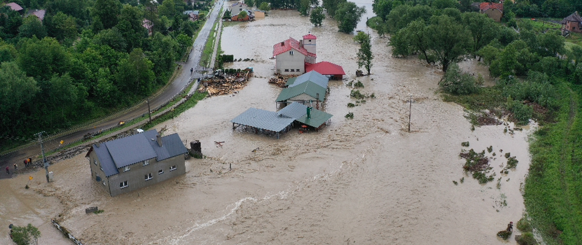 Zdjęcie z lotu ptaka przedstawia zalane budynki w gminie Jawornik Polski. Z lewej strony kadru widoczna jest droga do której dochodzi fala powodziowa. Po prawej stronie drogi widoczne cztery zalane budynki. Woda osiąga głębokość około jednego metra. Wyraźnie widoczne spiętrzenia, które sugerują dużą szybkość przemieszczania się wody. Woda jest brudna i mętna.