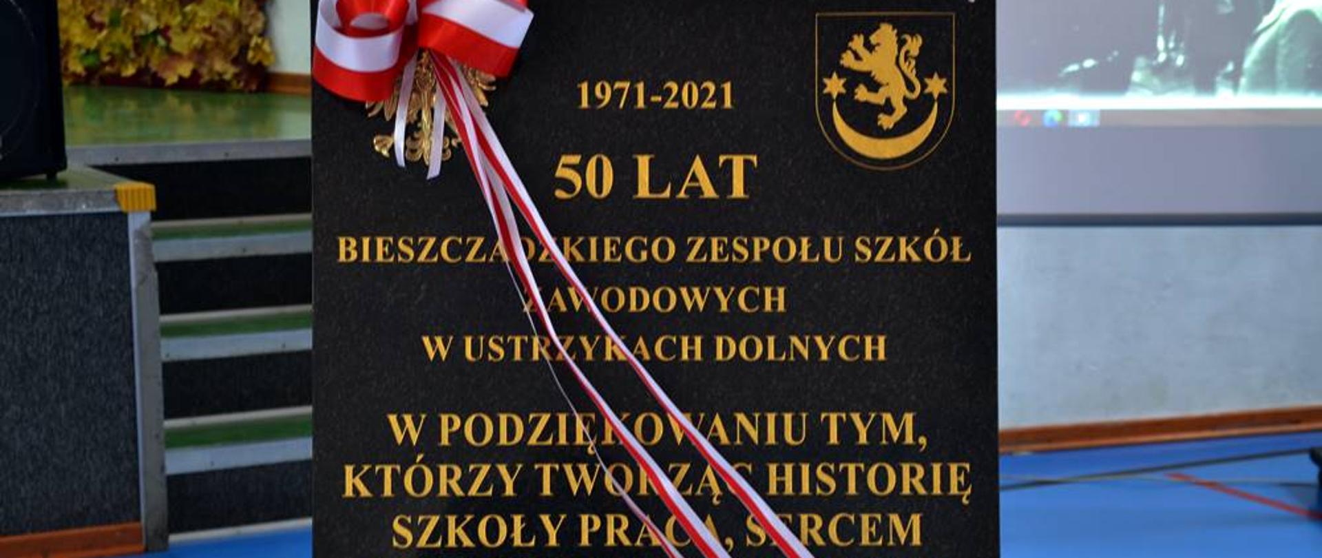Na zdjęciu widać tablicę pamiątkową z okazji 50 lecia Bieszczadzkiego Zespołu Szkół Zawodowych w Ustrzykach Dolnych. Tablica jest czarna i jest na niej złoty orzeł oraz herb powiatu oraz złoty napis "1971-2021 50 lat Bieszczadzkiego Zespołu Szkół Zawodowych w Ustrzykach Dolnych W podziękowaniu tym, którzy tworzą historię szkoły pracą, sercem i talentem przyczynili się do jej rozwoju Ustrzyki Dolne 13 października" Przez tablicę jest przewiązana biało-czerwona wstążka. 