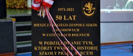 Na zdjęciu widać tablicę pamiątkową z okazji 50 lecia Bieszczadzkiego Zespołu Szkół Zawodowych w Ustrzykach Dolnych. Tablica jest czarna i jest na niej złoty orzeł oraz herb powiatu oraz złoty napis "1971-2021 50 lat Bieszczadzkiego Zespołu Szkół Zawodowych w Ustrzykach Dolnych W podziękowaniu tym, którzy tworzą historię szkoły pracą, sercem i talentem przyczynili się do jej rozwoju Ustrzyki Dolne 13 października" Przez tablicę jest przewiązana biało-czerwona wstążka. 