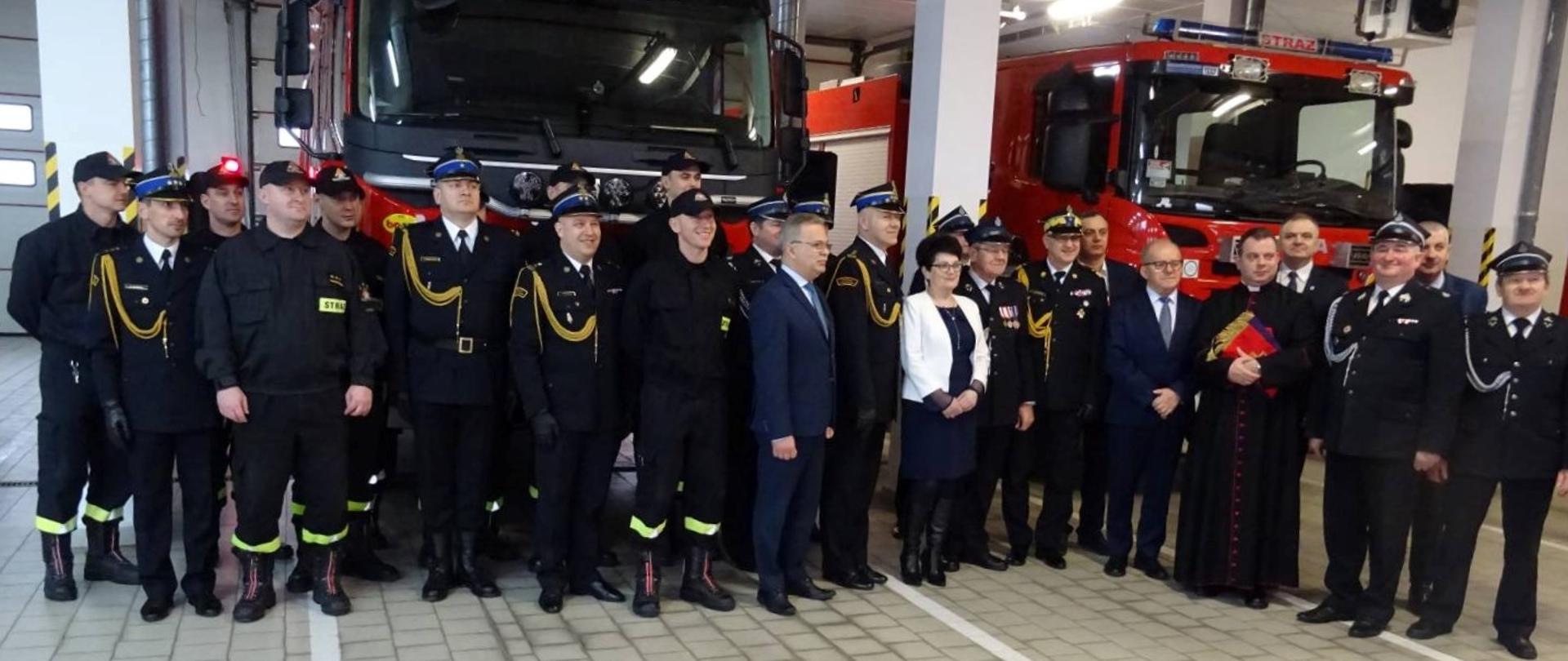 Grupowe zdjęcie gości uroczystości - ok. 20 osób, mundurowych i cywilów. W tle dwa wozy bojowe na swoich stanowiskach garażowych.