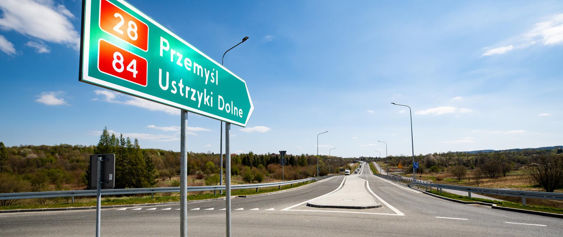 Na pierwszym planie znak drogowy wskazujący kierunek Przemyśl i Ustrzyki. Świeci słońce, jest pogodnie. Widać drogę i jadące w tle samochody. Zdjęcie zrobione z perspektywy ronda. 