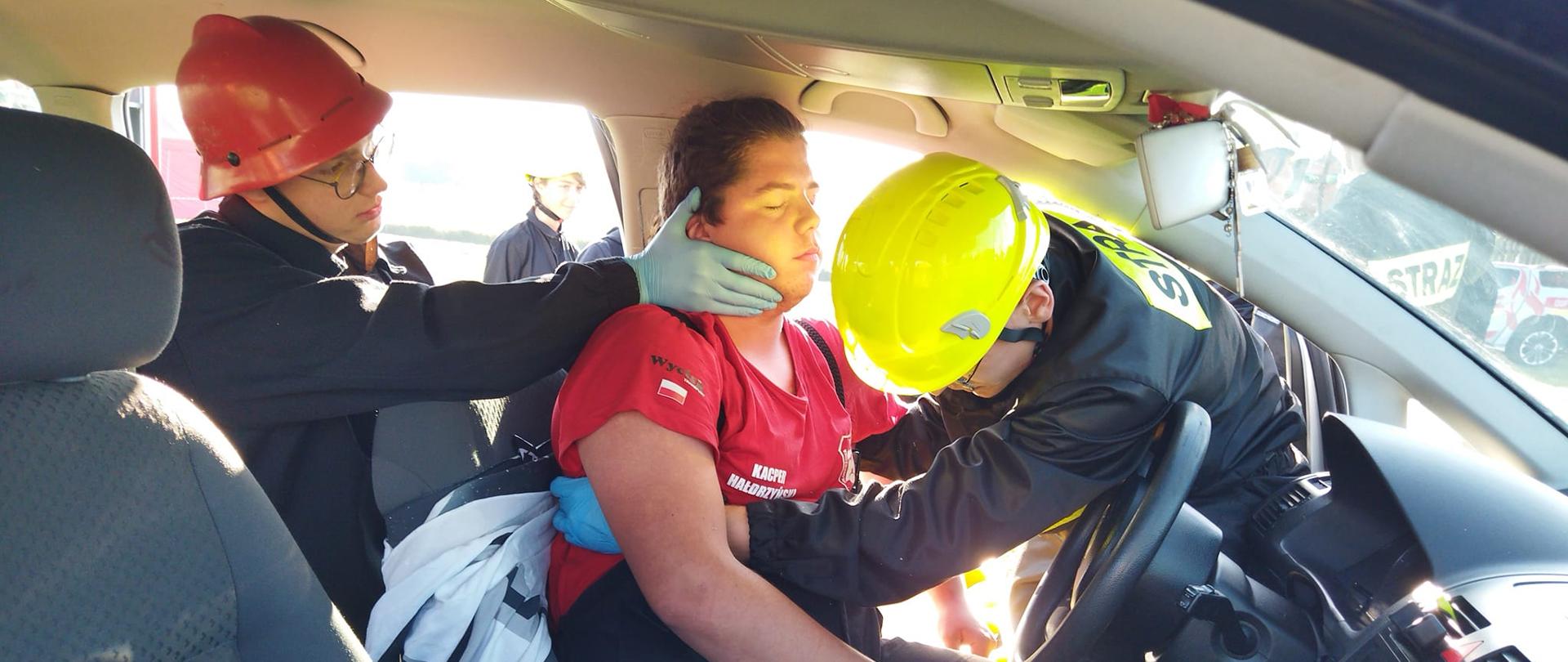 Obóz survivalowy w Sobiałkowie. Zdjęcie przedstawia wnętrze samochodu osobowego, w którym znajduje się "poszkodowany kierowca" oraz dwóch ratowników (uczestników obozu) udzielających mu pomocy. Jedez nich (w czerwonym hełmie) stabilizuje głowę, drugi (w żółtym hełmie) trzyma kierowcę pod ramionami. 