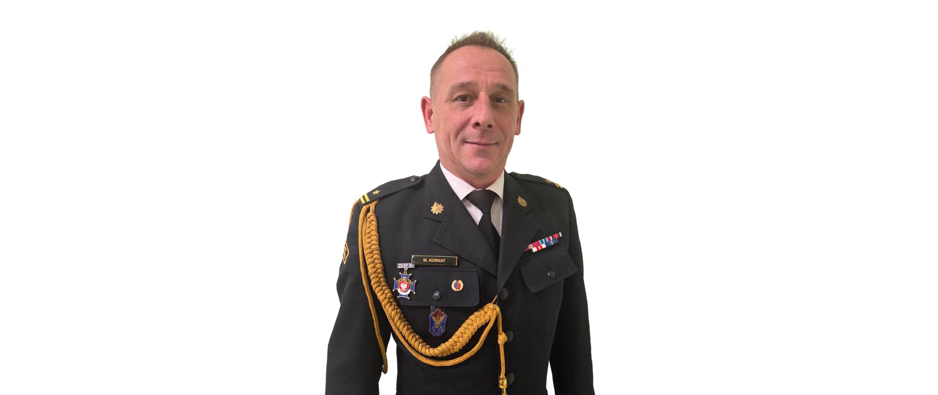 Zdjęcie przedstawia komendanta powiatowego Państwowej Straży Pożarnej w Łęczycy młodszego brygadiera Marcina Kornata w czarnym mundurze, na którym znajduje się stopień służbowy oraz odznaczenia strażackie. Zdjęcie portretowe na białym tle.