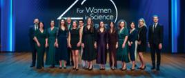 Na tle granatowej ściany z napisem 25 for Women in Science stoi kilkanaście osób, głównie elegancko ubranych kobiet.