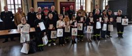 Grupa dzieci i młodzieży stoi w rzędzie w dużej, jasnej sali z wysokimi oknami i zasłonami. Uczestnicy trzymają dyplomy oraz torby z nagrodami, a część z nich ma elementy stroju strażackiego. Za nimi stoją dorośli, w tym osoby w mundurach strażackich. W tle widoczne są banery Ochotniczej i Państwowej Straży Pożarnej, co wskazuje na uroczyste wręczenie nagród w konkursie związanym ze strażą pożarną.