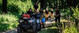 Zdjęcie przedstawia ćwiczenia strażackie w lesie. Na pierwszym planie, na gruntowej drodze leśnej, stoi duży, ciemnoszary quad ratowniczy z przyczepką. Pojazd wyposażony jest w oświetlenie LED oraz pomarańczową skrzynię sprzętową na przednim bagażniku. Za kierownicą quada siedzi strażak w czarnej koszulce i jaskrawozielonym kasku ochronnym. Na przyczepce oraz tuż za nią znajdują się inni strażacy w piaskowych spodniach mundurowych i czarnych koszulkach z elementami odblaskowymi, którzy rozwijają i asekurują długi, biały wąż strażacki ciągnący się wzdłuż drogi. Akcja rozgrywa się w pełnym słońcu, w otoczeniu gęstych drzew i zielonej roślinności. W prawym dolnym rogu widnieje podpis: „st. ogn. Marcin Herok | Państwowa Straż Pożarna”.