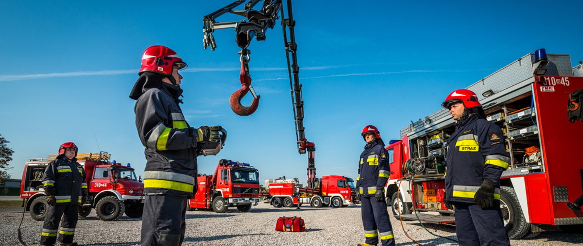 Strażacy stojący wśród sprzętu ratownictwa technicznego. W tle samochody ratownicze - jeden z nich z wysuniętym ramieniem dźwigu i hakiem.