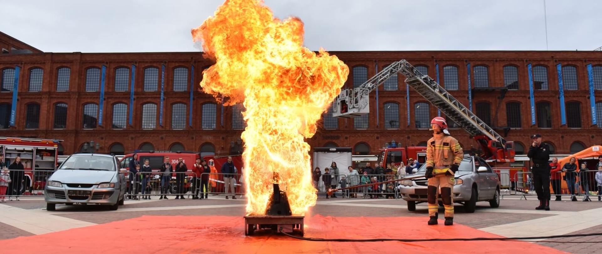 na głównym planie słup ognia nad metalowym urządzeniem obok strażak w mundurze bojowym w tle 2 wraki samochodów ludzie, samochody strażackie oraz budynek