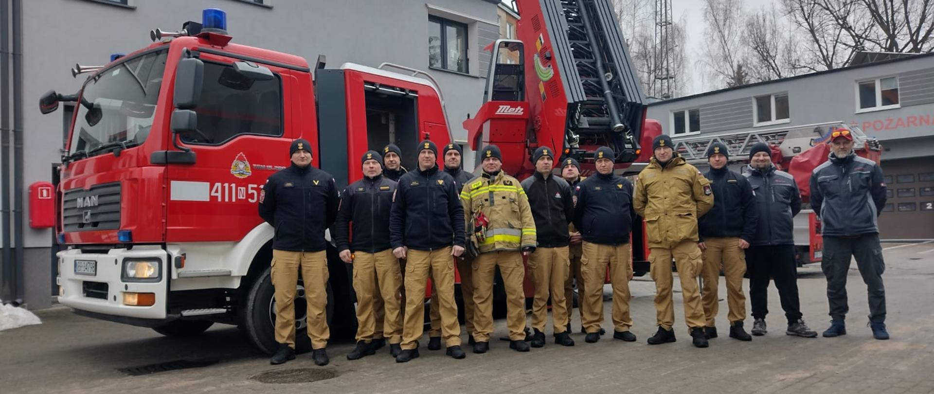 Na zdjęciu grupa strażaków, stoją obok siebie. W tal autodrabina strażacka.