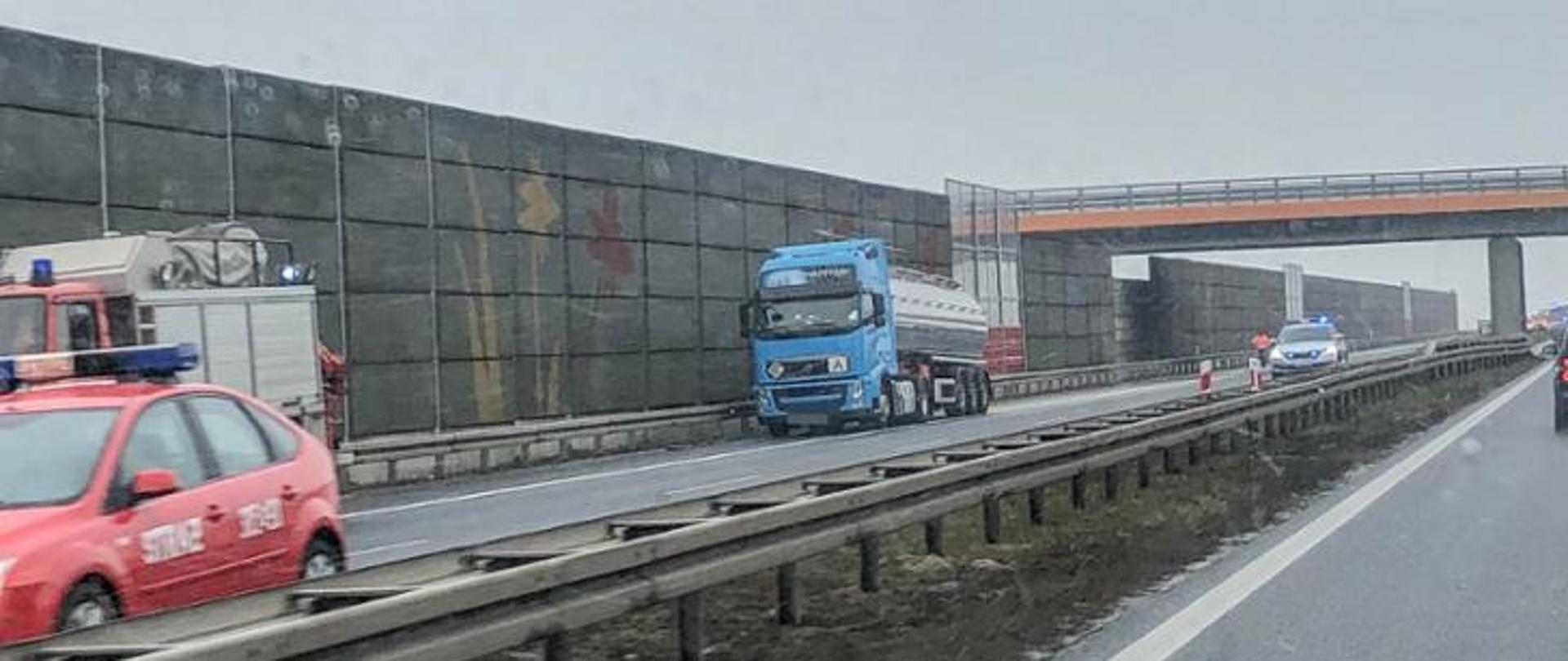 Zdjęcie zrobione na drodze szybkiego ruchu. Na zdjęciu cztery samochody. Po prawej stronie samochód typu TIR oraz samochód policji z wałczonymi niebieskimi światłami uprzywilejowania . Po lewej stronie dwa samochody straży pożarnej. Na zdjęciu również wiadukt nad drogą szybkiego ruchu oraz ekrany dźwiękochłonne