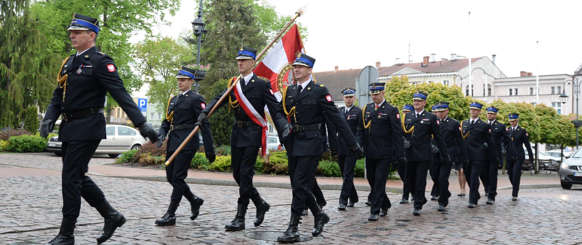 1. Uroczysty przemarsz strażaków w czarnych mundurach galowych po brukowanym placu. Na czele idzie dowódca, za nim maszeruje poczet sztandarowy niosący biało-czerwoną flagę z godłem, a w tle widać kolejnych strażaków w szyku, zieleń drzew oraz miejską zabudowę.