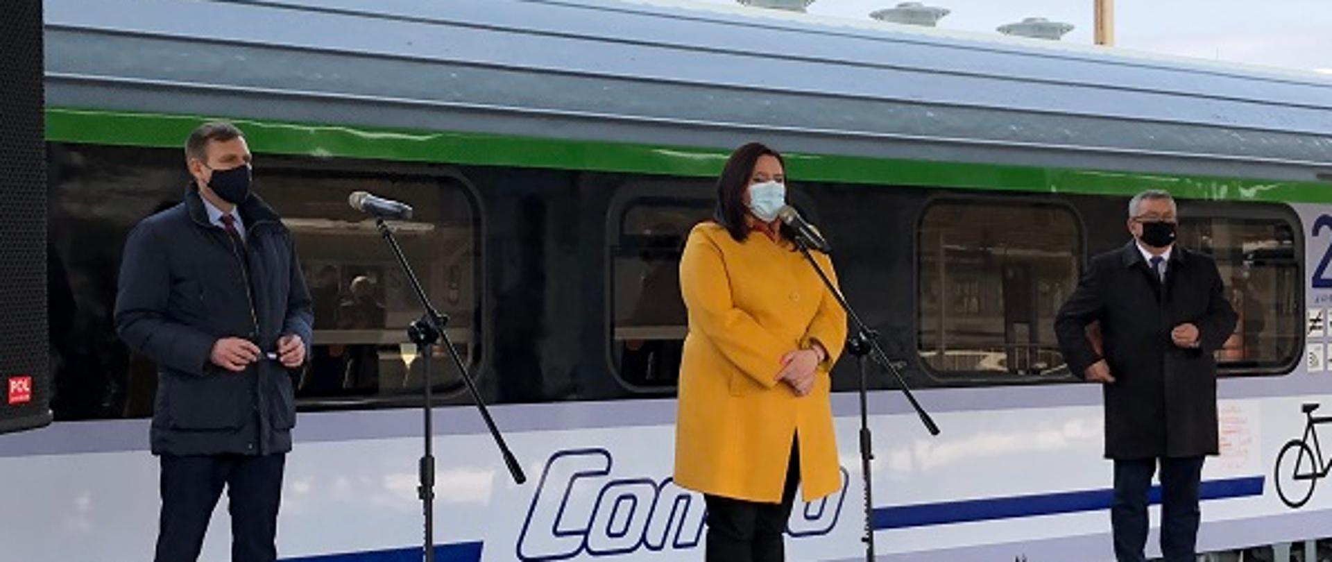 Na pierwszym planie trzy osoby stoją przed mikrofonami na podeście. W środku minister Małgorzata Jarosińska-Jedynak.