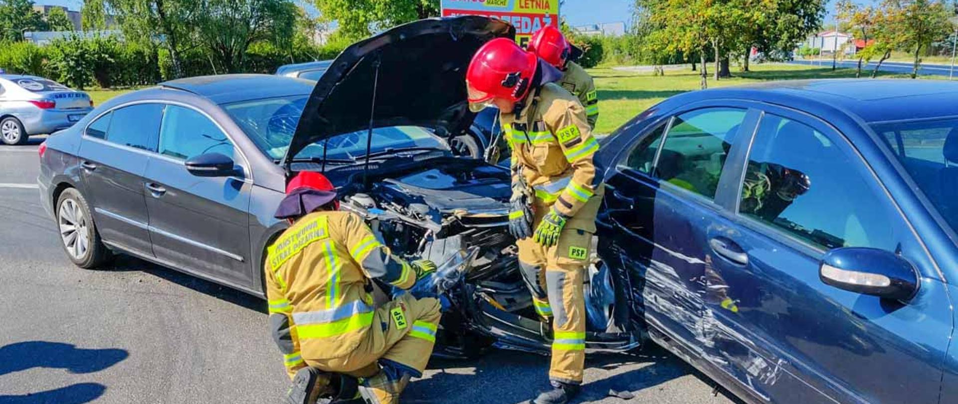 Na zdjęciu 2 uszkodzone pojazdy przy nich dwóch strażaków w ubraniach specjalnych podczas czynności ratowniczych