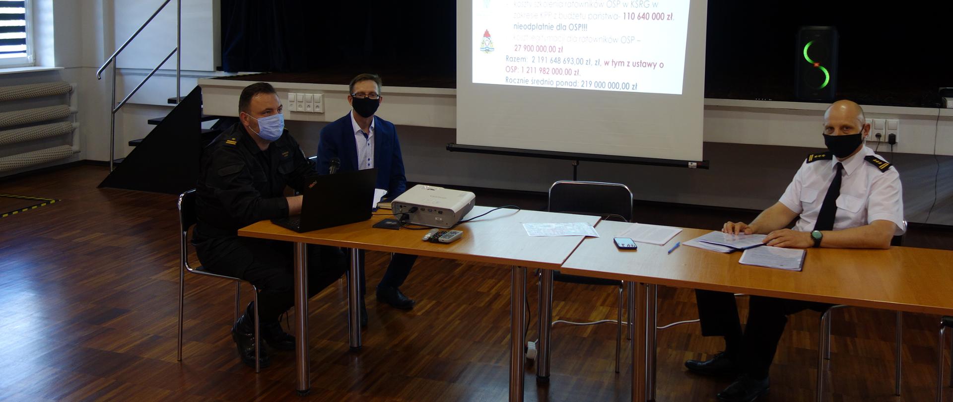 W sali OSP przy stole siedzą Komendant Powiatowy Państwowej Straży Pożarnej w Kutnie wraz z naczelnikiem i przedstawicielem OSP. Na stole widać dokumenty i rzutnik multimedialny, który na makiecie wyświetla informacje.