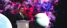 Widok z boku pod kątem, za mównicą stoi minister Kulasek i mówi do mikrofonu, za nim wielki ekran z obrazem kilku planet na czarno-fioletowym tle.