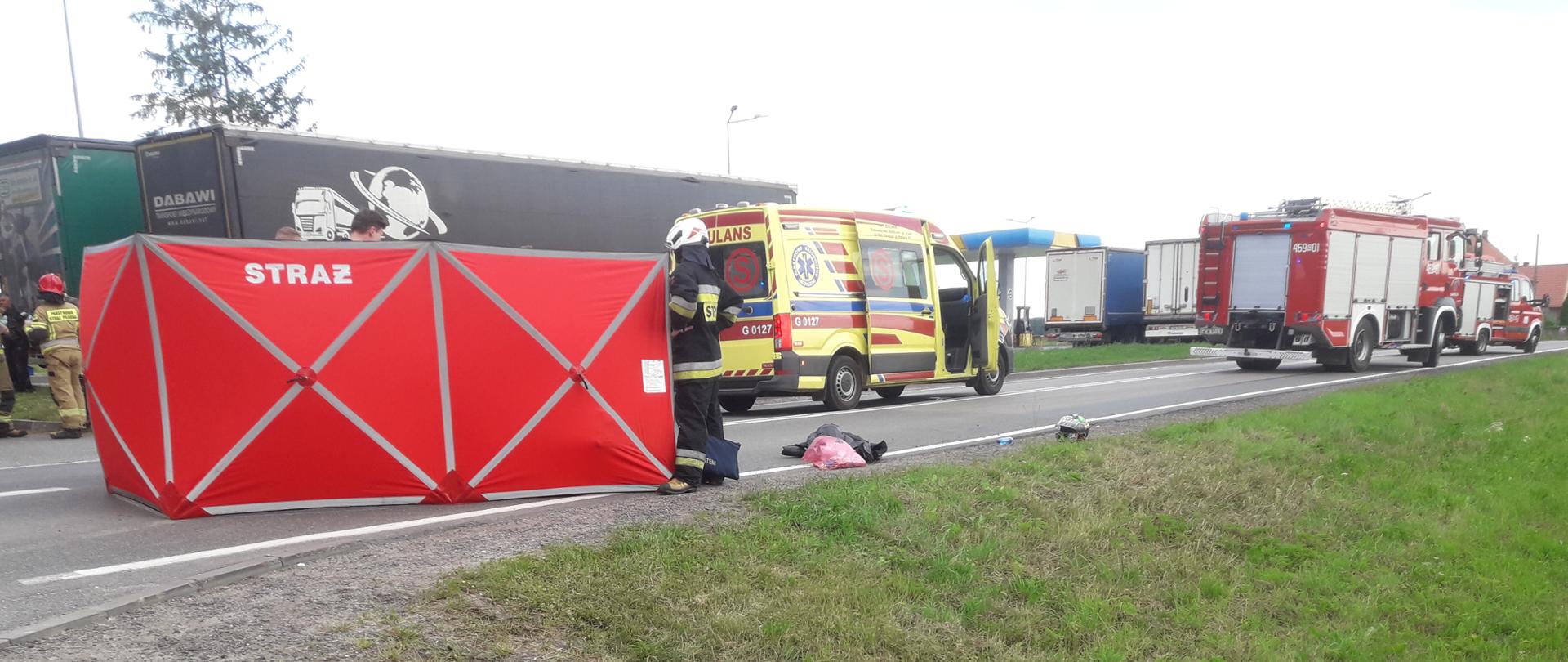 Miejsce zdarzenia osłonięte parawanem, obok motocykl, w tle pojazdy podmiotów ratowniczych
