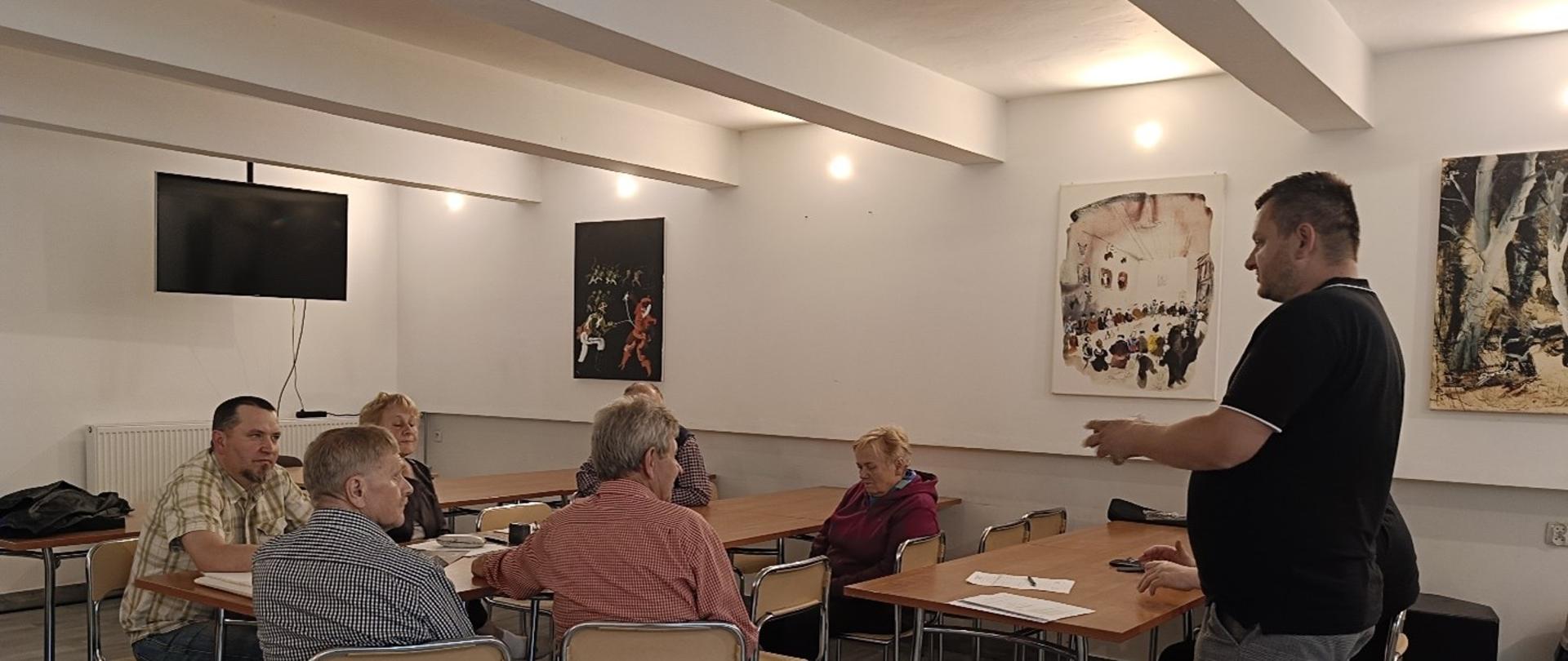 Grupa ludzi siedząca przy jasnym stole. Jeden mężczyzna stoi po prawej stronie. Pomieszczenie jest jasne. Ze ściany zwisa telewizor. Na ścianach wiszą obrazy.