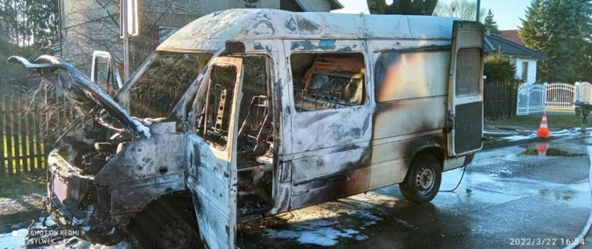 Zdjęcie wykonane w dzień. Zdjęcie przedstawia spalone auto dostawcze z boku. W tle domki jednorodzinne. Auto ma podniesioną maskę, otwarte boczne i tyle drzwi. Pod autem dużo piany.
