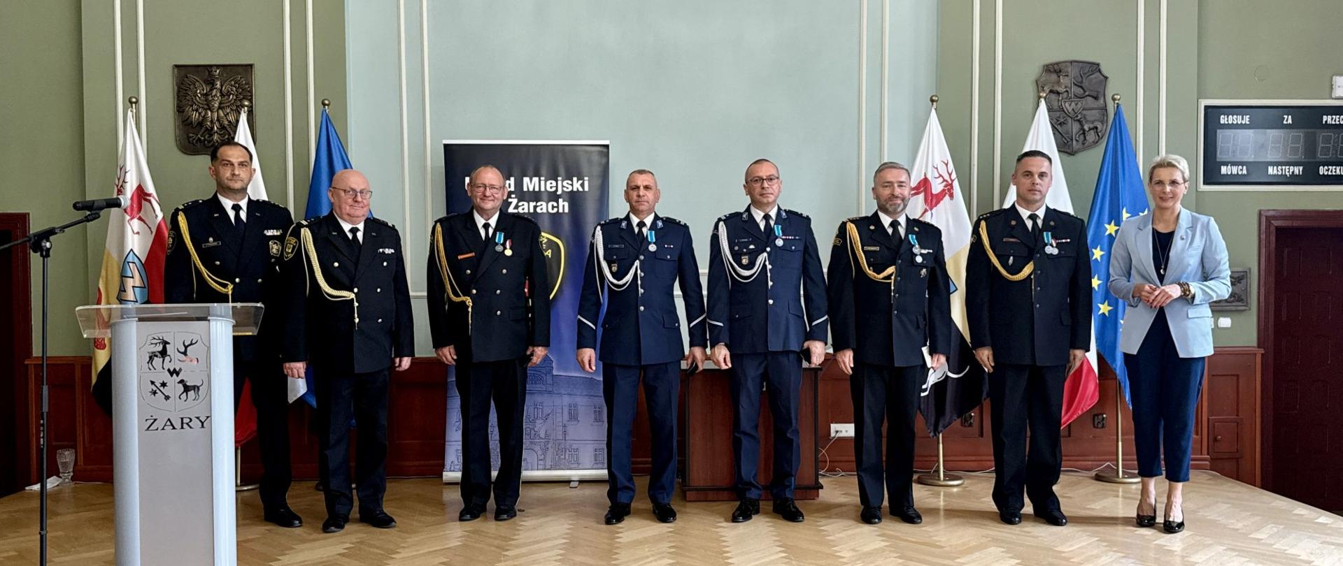 Pamiątkowa fotografia wykonana na drewnianym podwyższeniu w sali ratusza. W rzędzie stoi siedmiu umundurowanych funkcjonariuszy (w tym Komendant Powiatowy PSP st. bryg. Paweł Hryniewicz i Zastępca Komendanta Powiatowego PSP kpt. Paweł Brela z medalami) oraz po prawej stronie Burmistrz Edyta Gajda. Po lewej stronie widoczna jest mównica z herbem Żar, a w tle rząd flag i baner Urzędu Miejskiego.