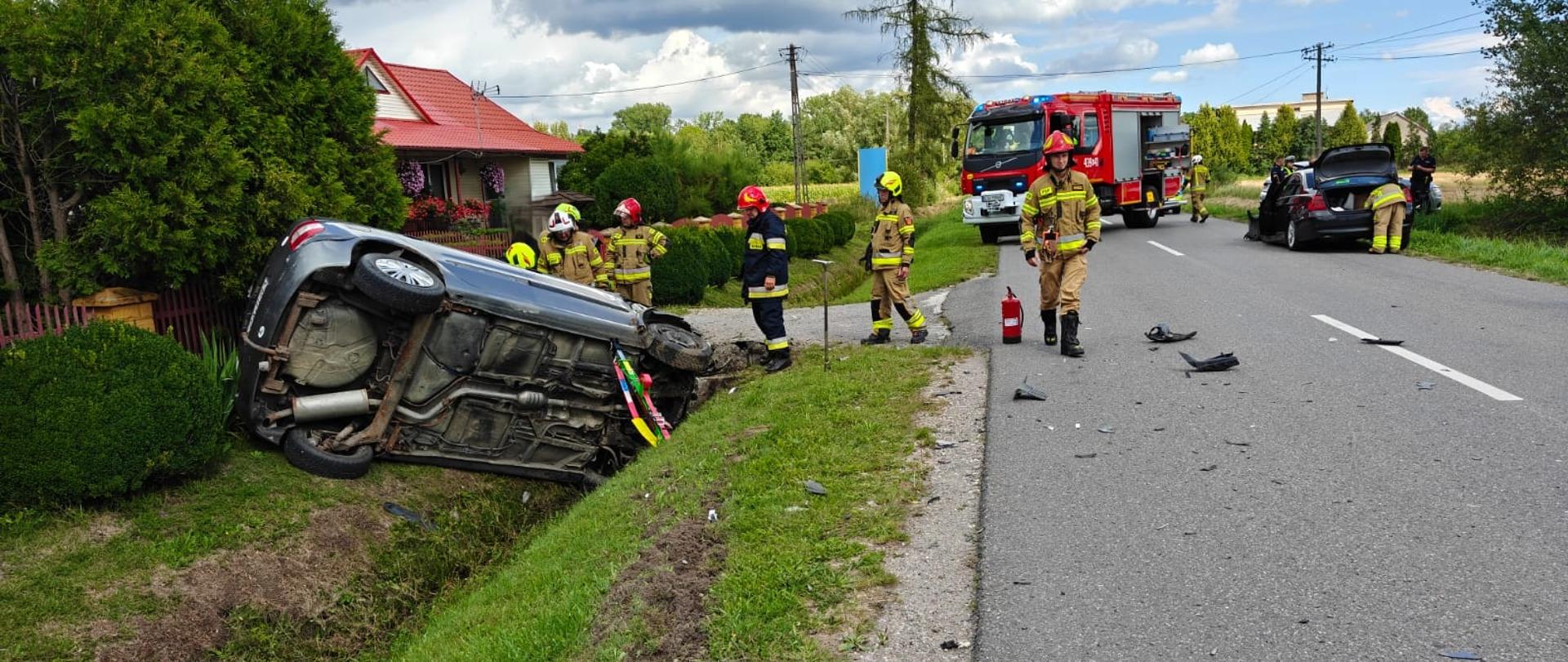 Na zdjęciu widać jeden z pojazdów który brał udział w wypadku i uderzył w mostek posesji, auto na lewym boku zabezpieczone podporą ratowniczą przez strażaków, w oddali pojazd ratowniczy strażaków i drugi pojazd bioracy udział w zdarzeniu, na drodze plastiki po wypadku oraz strażacy podczas prowadzenia swoich działań.