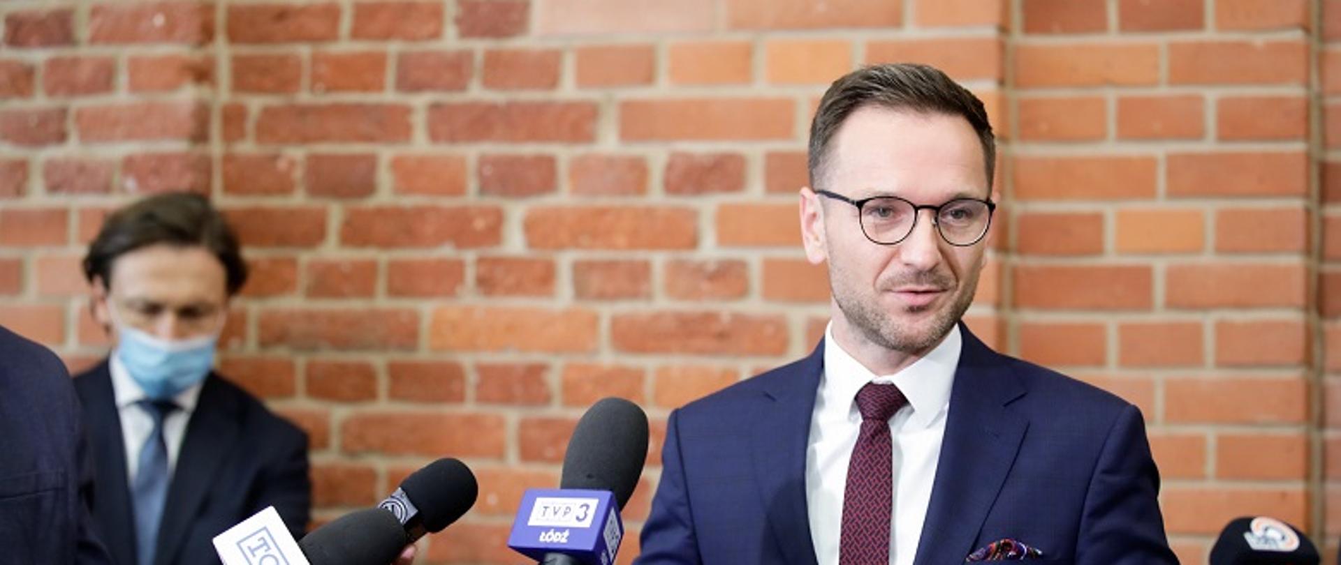 Minister Waldemar Buda podczas konferencji prasowej, stoi, a przed nim widoczne mikrofony dziennikarzy