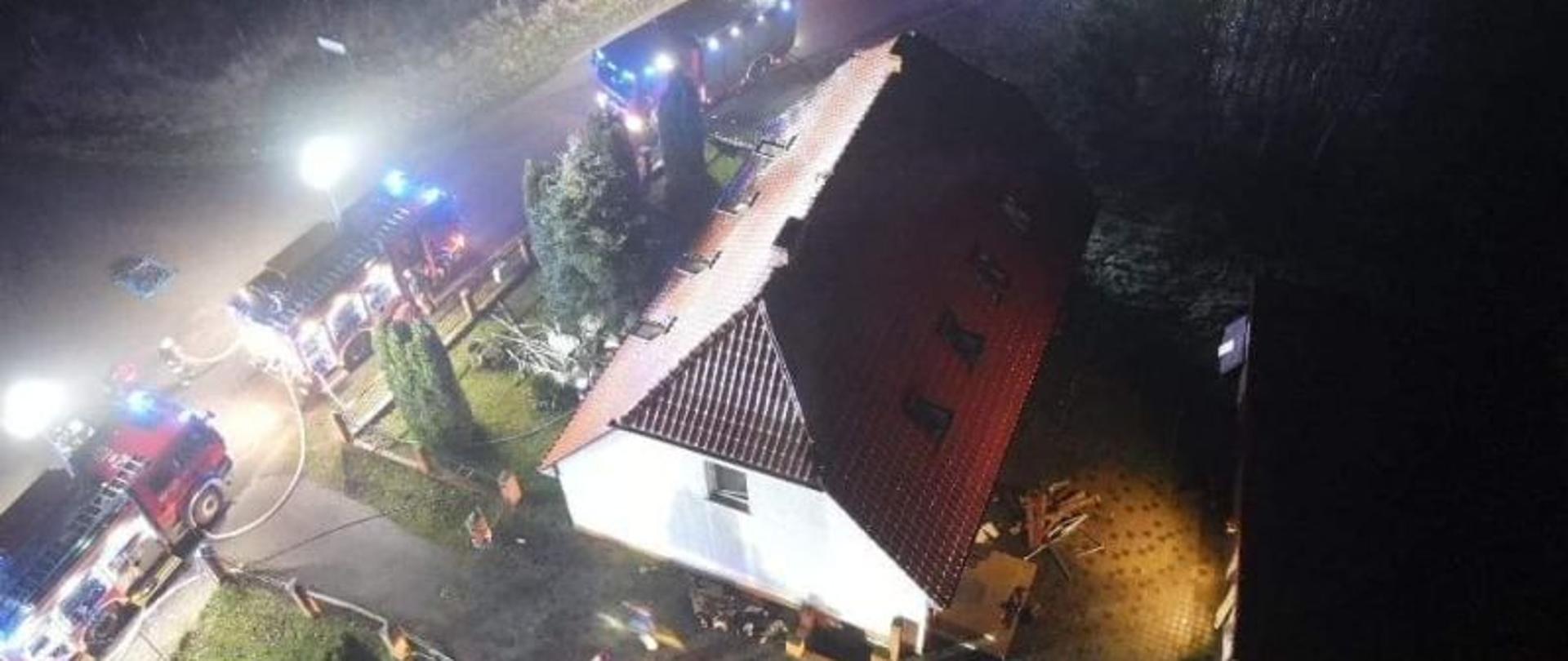 Widok z góry, z drona na miejsce prowadzenia działań. Centralny punkt zdjęcia stanowi dom. Od góry droga wzdłuż, której stoją auta strażackie. Teren oświetlają najaśnice strażackie.