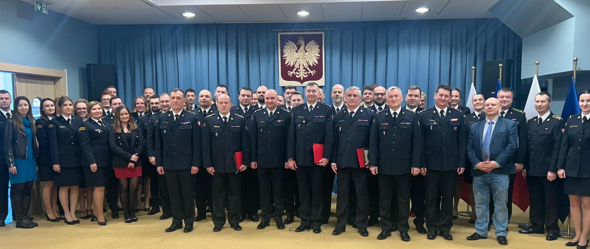 Zdjęcie grupowe uczestników uroczystości na tle niebieskiej ściany na której wisi godło Polski. 