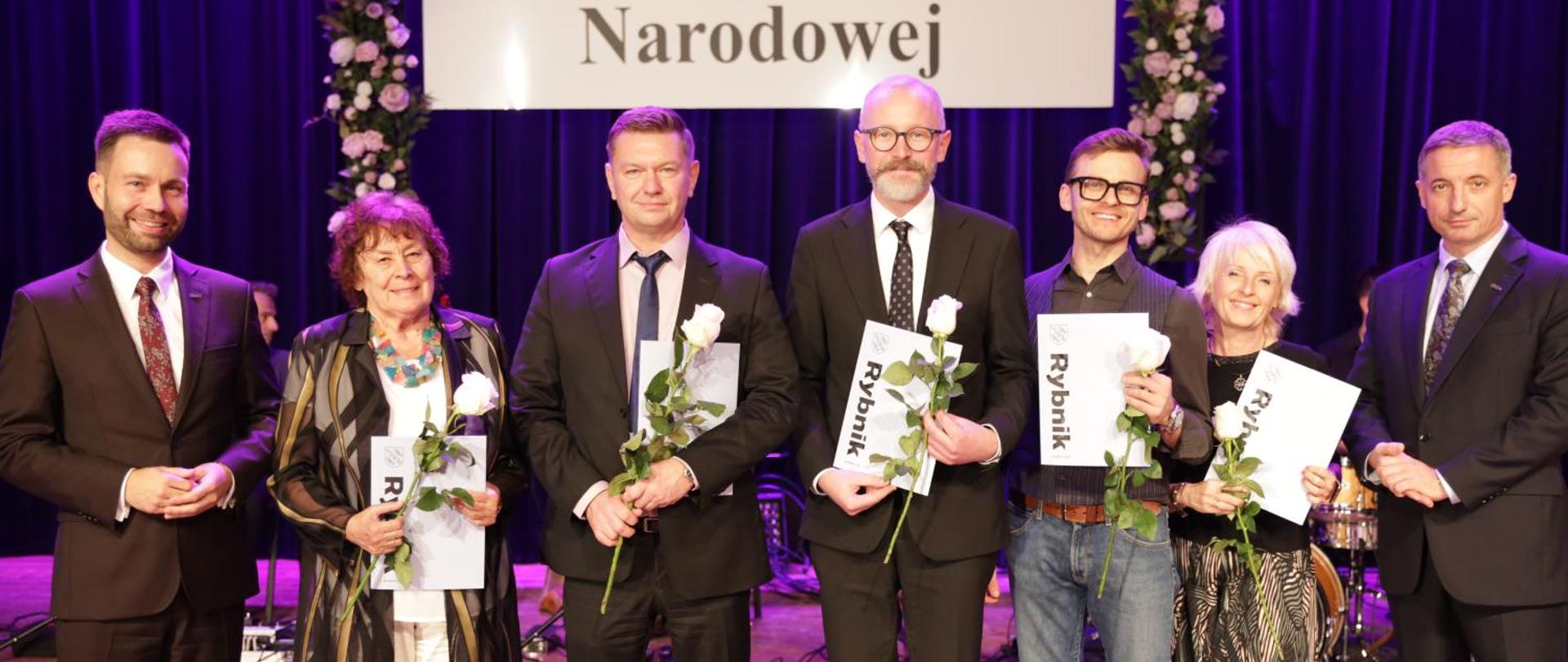 Kolorowe zdjęcie, na którym stoi w jednym rzędzie 7 osób tj. Prezydenci Miasta Rybnika (po bokach, z prawej i lewej strony), a między nimi nagrodzeni nauczyciele naszej szkoły, którzy trzymają w rękach niebieskie teczki z listami gratulacyjnymi i białe róże. Wszyscy uśmiechnięci i ubrani w eleganckie stroje. W tle granatowa kurtyna z dekoracją kwiatową i białą tablicą z czarnym napisem Dzień Edukacji Narodowej.