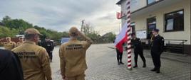 Na zdjęciu widoczny na placu zewnętrznym przed Komendą PSP w Gorlicach poczet flagowy przy maszcie w ubraniach reprezentacyjnych podczas podnoszenia flagi w zawiązku z uroczystością Dnia Flagi Rzeczypospolitej Polskiej. Na zdjęciu widoczne również zmiany służbowe w dwuszeregu w pozycji na baczność w ubraniach koszarowych koloru pisakowego i czarnego w czapkach.