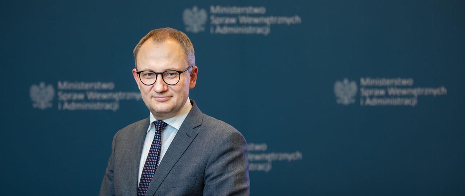 Wiceminister Błażej Poboży na tle granatowej ścianki z logiem Ministerstwa Spraw Wewnętrznych i Administracji.