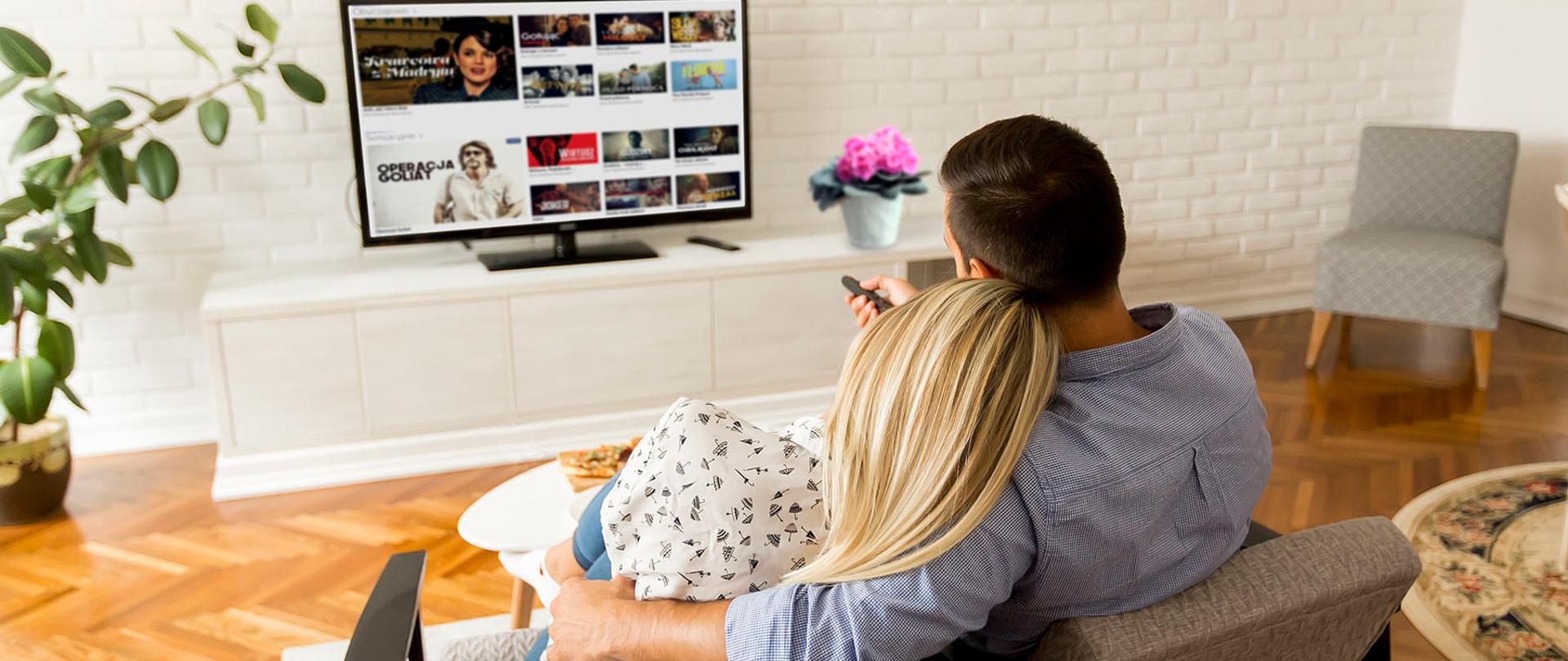 Zdjęcie ilustracyjne w poziomie. Widok z tyłu pary oglądającej telewizję w salonie