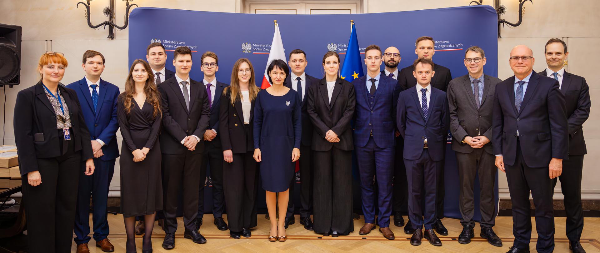 Na zdjęciu stoi grupa osób. W tle ścianka z logotypem MSZ.