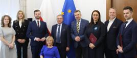 Pod ścianą stoi grupa osób trzymających czerwone teczki, pośrodku grupy minister Wieczorek, za nimi pod ścianą flagi Polski i UE.
