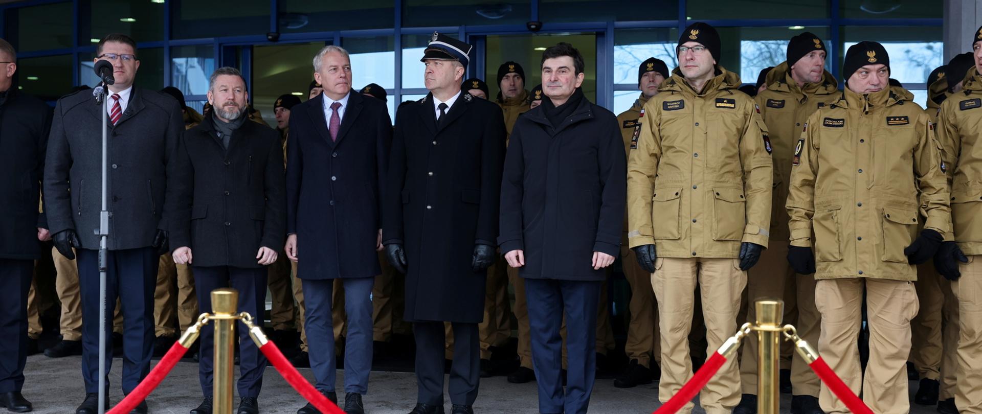 Grupa funkcjonariuszy Państwowej Straży Pożarnej oraz przedstawicieli administracji publicznej stoi przed budynkiem podczas oficjalnej części uroczystości