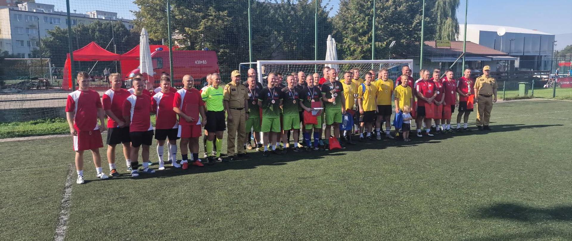 Na boisku piłkarskim, przed bramką, stoją 4 drużyny piłkarskie wraz z Komendantem Wojewódzkim PSP i jego Zastępcą, a także sędziowie w zielonych koszulkach.