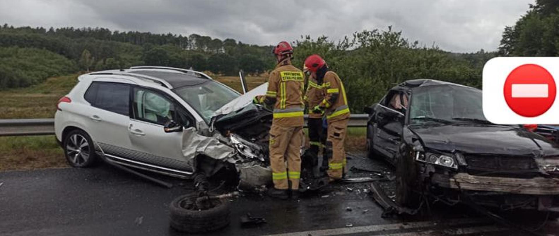 Widok na dwa rozbite auta. Po lewej białe z rozbitym przodem a po prawej czarne z rozbitym bokiem. Po środku dwa strażacy prowadzący swoje czynności. Tło stanowi pochmurne niebo.