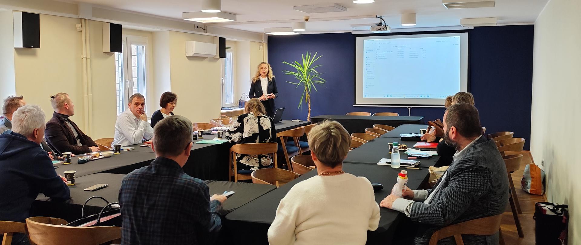 W przestronnej, nowocześnie urządzonej sali konferencyjnej trwa spotkanie szkoleniowe. Uczestnicy siedzą przy stołach ustawionych w kształt litery U, zwróceni w stronę Wojewódzkiej Inspektor prowadzącej spotkanie, która stoi obok ekranu projekcyjnego. Część osób robi notatki, inni słuchają lub trzymają w dłoniach materiały szkoleniowe i kubki z napojami.