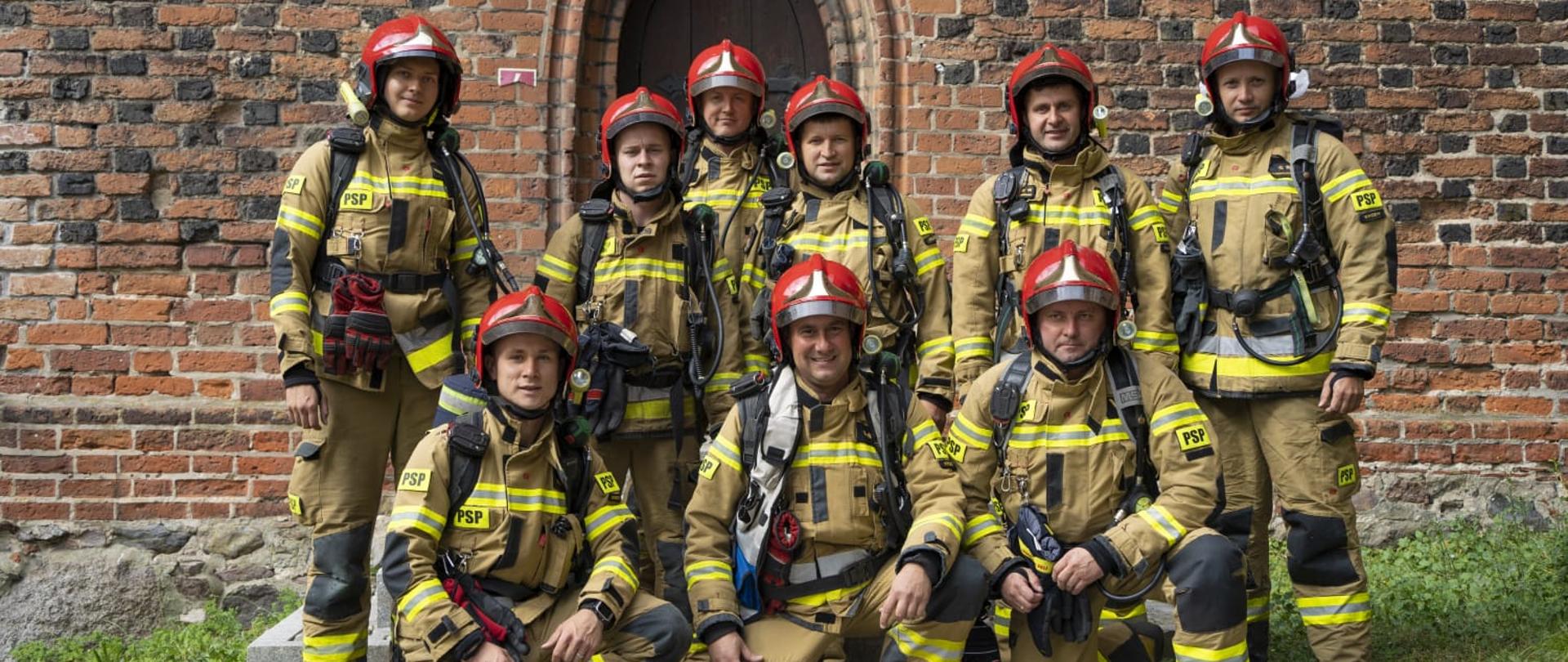 Grupa strażaków w ubraniach bojowych pozująca do zdjęcia przed wejściem do wieży.