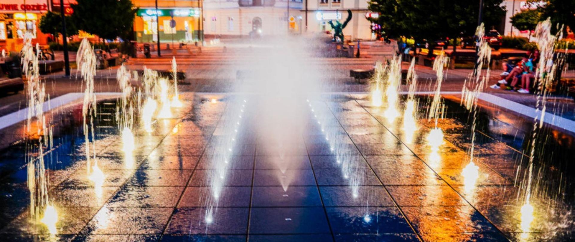 Zdjęcie przedstawia fontannę na rynku mieleckim, wieczorową porą. Fontanny podświetlone diodami LED.