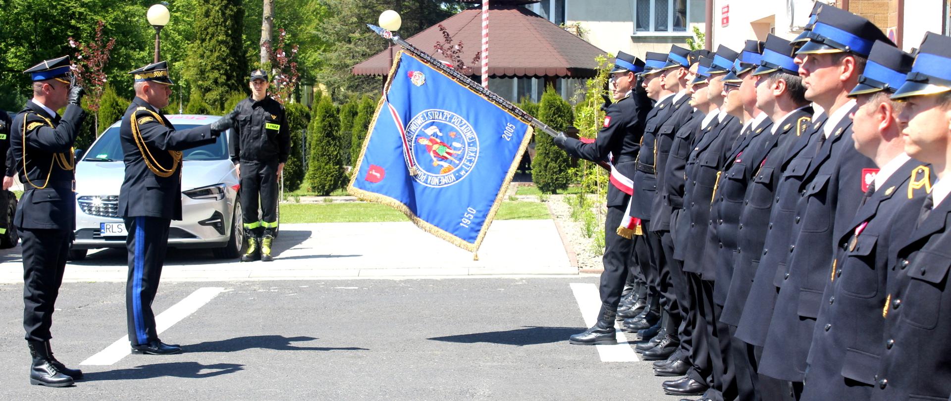 Zdjęcie wykonane na zewnątrz. Po prawej stronie stoją strażacy ubrani w mundury wyjściowe, ustawieni w szeregu. Jeden ze strażaków trzyma pochylony do przodu sztandar Komendy Powiatowej Państwowej Straży Pożarnej. Po lewej stronie zdjęcia dwóch oficerów Państwowej Straży Pożarnej salutuje sztandarowi. Jeden z oficerów jest w stopniu generała.