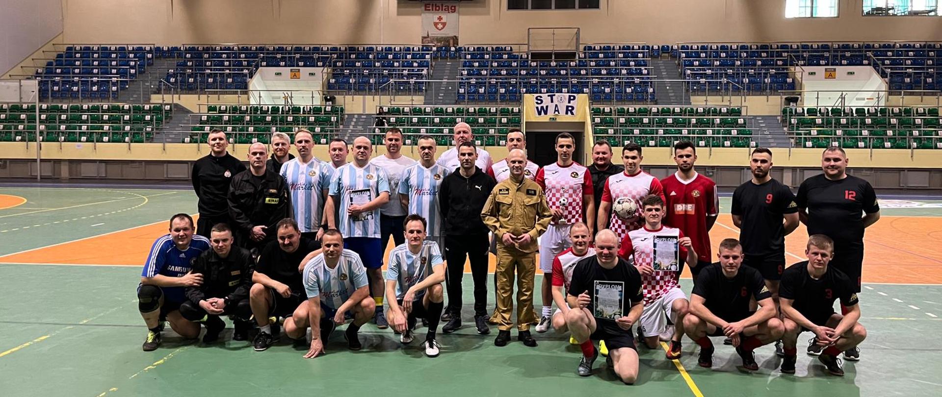 Grupowe zdjęcie strażaków w strojach sportowych na płycie boiska. W tle widoczne trybuny.