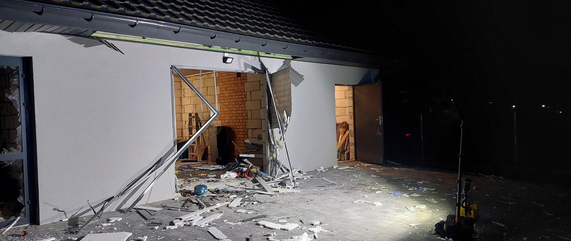 Zdjęcie przedstawia fasadę budynku gospodarczego po wybuchu. Na zdjęciu widoczne są zniszczenia wywołane eksplozją, w tym otwór w którym byla brama garażowa. Elewacja budynku koloru szarego. Noc.