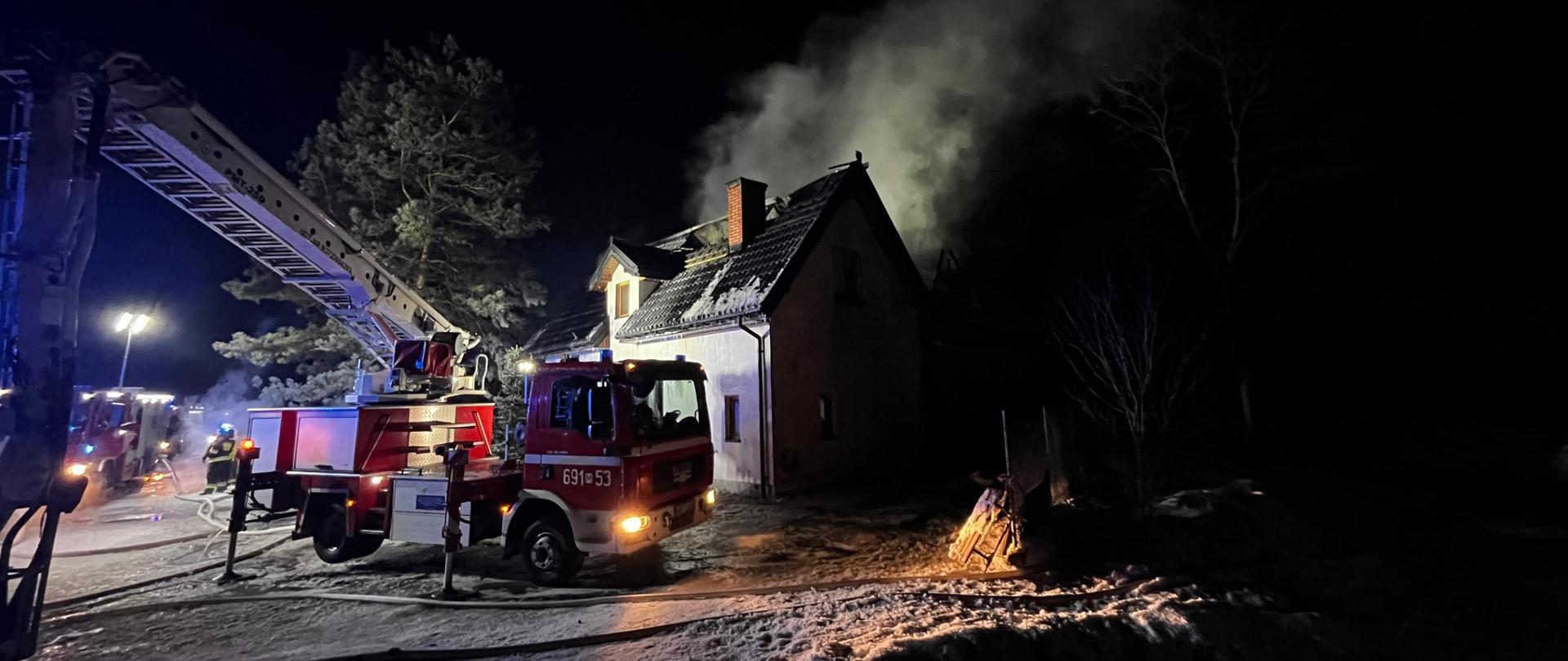 Rozstawiony podnośnik strażacki stoi przed domem, z którego leci dym. Pora nocna.