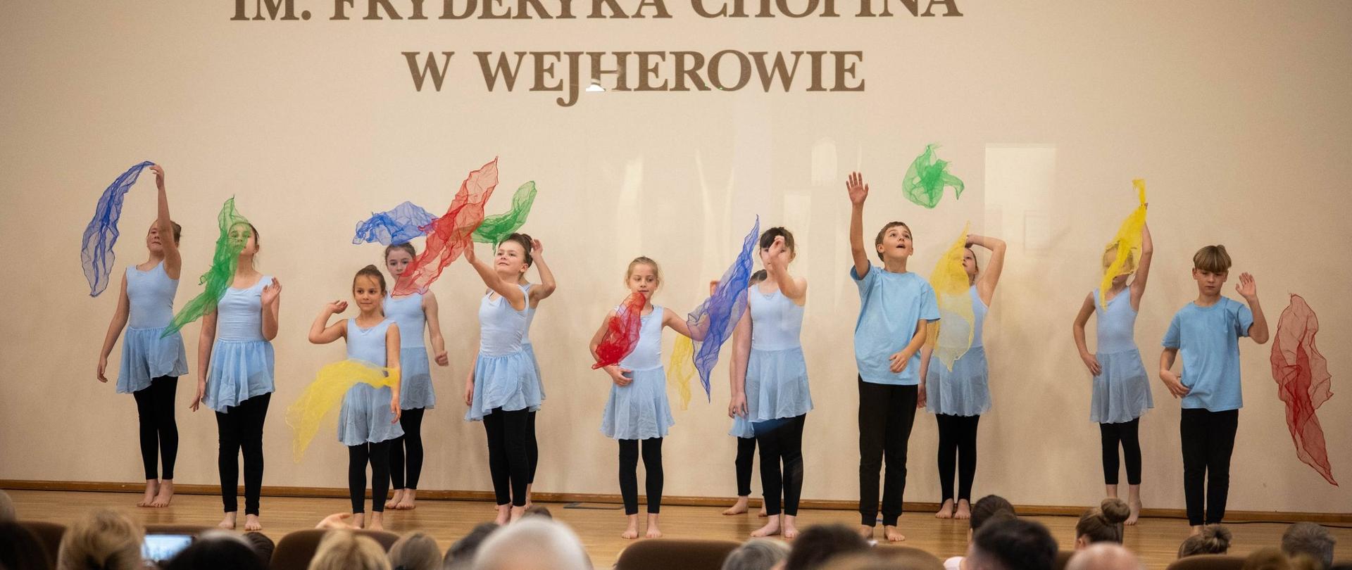 Na zdjęciu dzieci w niebieskich strojach tańczą wraz z kolorowymi chustami. Duża dynamika fotografii