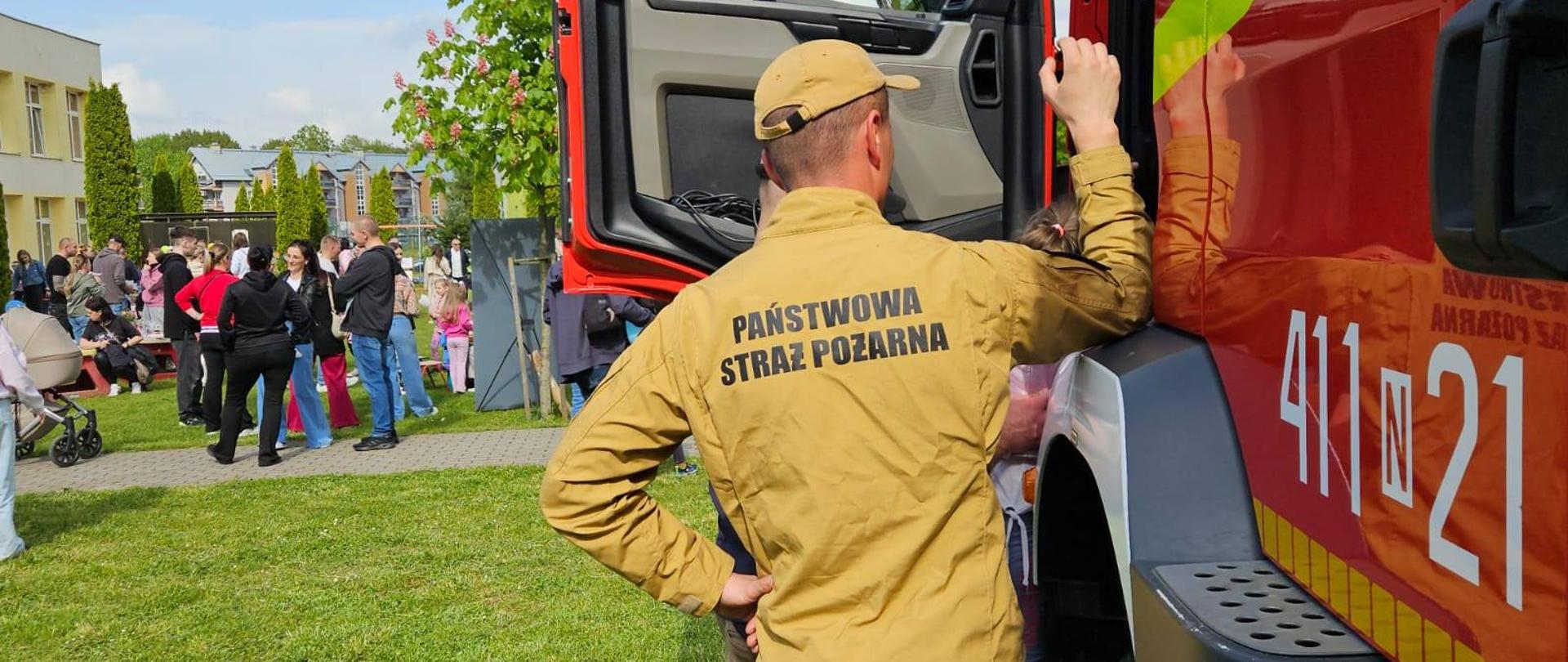 Teren przedszkola. Z prawej strony czerwony samochód strażacki, strażak stoi przy samochodzie, dzieci wchodzą do samochodu.Z lewej strony rodzice z dziećmi. Zielona trawa.